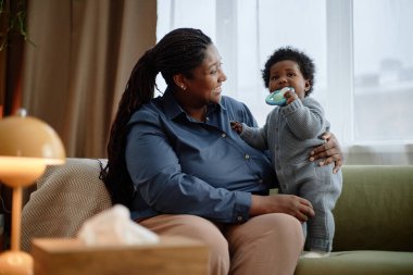 Orta boy gülümseyen siyah bir kadın kucağında küçük bir çocukla yatak odasındaki kanepede oturmuş kızına bakarken bebek ayağa kalkıp diş çıkarma oyuncağını ısırıyor.