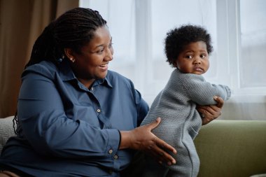 Orta derecede mutlu, siyah bir kadın, rahat örülmüş mavi bir elbise giymiş küçük bir kızı nazikçe kucağında tutuyor. Kanepede dikiliyor ve evdeki pencereye yaslanıyor.