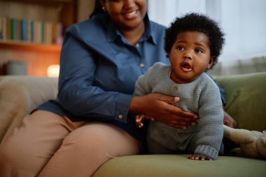 Meraklı küçük siyah kız rahat örülü tulumlar giyip emeklemeyi öğrenirken şefkatli anne kızını elleriyle destekliyor oturma odasındaki kanepede oturuyor.