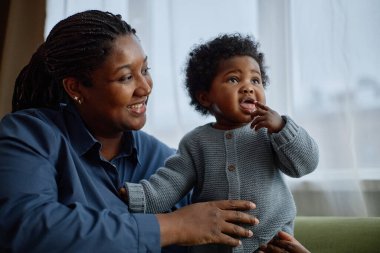 Orta pişmiş bir gülümseme. Siyah bir kadın, tatlı küçük bir kızı nazikçe kucağında tutuyor. Ağzı açık, mavi örülü bir şamata giyiyor.