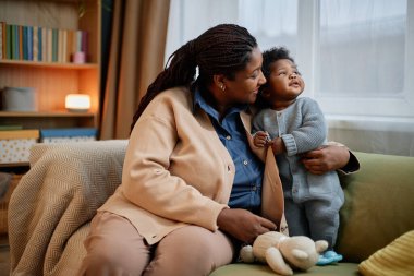 Orta ölçekli mutlu, rahat bir hırka giyen siyah bir kadın. Mavi örülü bebek kızı nazikçe kucaklıyor. Evdeki kanepede içtenlikle bağlanıyor.