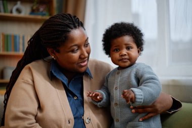 Siyahi bir kadının rahat bir hırka giyip, evde kanepede dinlenirken küçük kıza şefkatle baktığı, mavi örülmüş bir tulum giymiş kızına nazikçe sarıldığı bir fotoğraf.