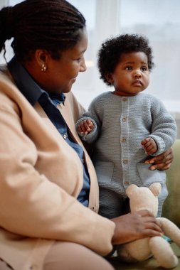 Siyah Anne 'nin bebeği nazikçe kucağına aldığı dikey fotoğraf. Kızı kanepede dikilirken. Evde birlikte biraz üzgün bir şekilde çekilmiş.