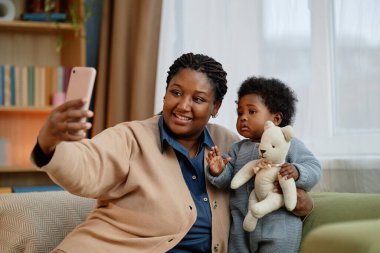 Küçük kızıyla selfie çekerken orta boy siyah bir kadın, oturma odasındaki kanepede el ele yumuşak oyuncaklar tutarken.