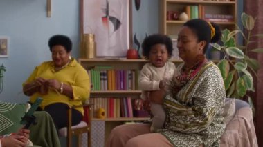 Akıllı telefonuyla oynayan genç bir kız, örgü ören bir kadın ve sıcak oturma odasında gülümseyen bir bebek tutan başka bir kadın.