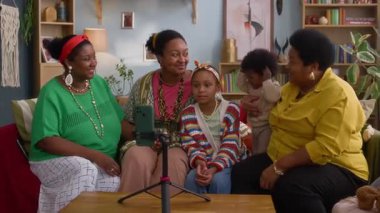 Renkli giysiler içinde Afrikalı Amerikalı bir grup tripodla akıllı telefona poz veriyor ve gülümsüyor, çizgili süveterli bir çocuk rahat oturma odasına uzanıyor.