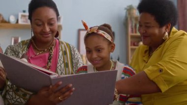 Üç kadın ve iki çocuklu neşeli Afro-Amerikan ailesi, büyük albümün etrafında toplandı, oturma odasında birlikte fotoğraflara baktılar.