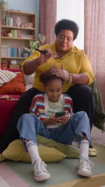 Torununun kıvırcık saçlarını tarayan Afrikalı Amerikalı büyükannesinin akıllı telefonuna odaklanmış dikey görüntüsü.