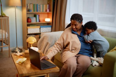 Genç siyahi kadın, kucuk kiz bebegiyle kanepede otururken kucuk bir dizüstü bilgisayarla kizinin rahat oturma odasinda annesine yaslandigi komik karikatürler izliyor.