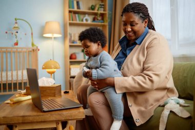 Orta halli mutlu siyah anne, kucağında küçük bir bebekle kanepede oturmuş dizüstü bilgisayar kullanarak komik çizgi filmler izliyor, rahat yatak odasındaki ahşap masaya yerleştiriliyor.