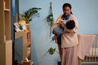 Siyahi anne, küçük bebeğini sevgiyle uyuturken, yorgun kızını kucağına alıp, yeşil bitkilerle sıcak çocukların yatak odasına koyarken orta boy bir fotoğraf.