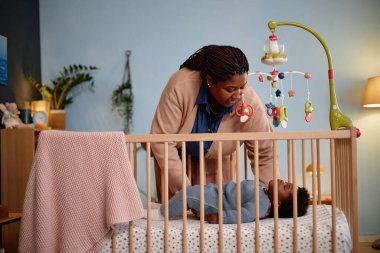 Önemseyen siyah anne, küçük kızını beşiğine koyuyor. Mavi pastel duvarlı, rahat yatak odasında uzanan yorgun, uykuya dalmış bebeğe bakıyor.