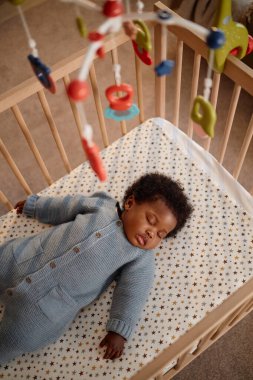 Mavi örülü tulum giymiş, bebek beşiğinde huzur içinde yatarken, müzik hareketleriyle yatarken, evde oturan çocuğun üzerinde asılı duran yatarken dikey yüksek açılı bir fotoğraf.