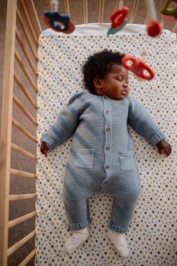 Dikey çekimde, mavi örülü romper giymiş küçük siyah bir kız bebek karyolasında cep telefonlarıyla yıldızlı battaniyede uyuyor, yatak odasındaki çocuğun üstünde asılı.