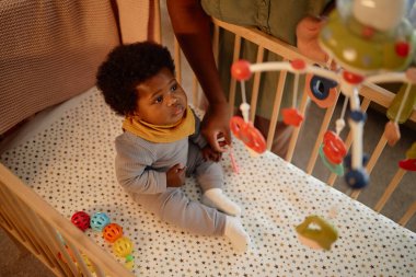 Yüksek açılı çekimde, yorgun siyah bebek yatakta oturur ve beşikteki hareketlere bakar. Annesi çocuğu sıcak bir ışıkla sıcak bir yatak odasında uyurken yukarıda asılı durur.