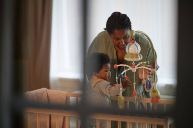 Siyah Anne 'nin bebek yatağının üzerine eğildiği orta boy bir fotoğraf. Küçük kız, yatak odasının penceresine yansıtılan müzikal bebek arabalarıyla oynarken. Huzurlu bir ev hayatı sahnesi.