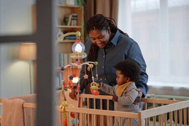 Orta ölçekli bir gülümseme. Siyah anne, küçük kızına şefkatle bakarken bebek de rahat yatak odasında hareketli beşiklerle oynuyor. Anneliğin mutlu anı.
