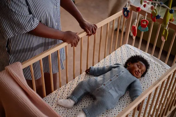 Mavi renkli örülü tulum giymiş küçük siyahi çocuğun yukarıda bebek arabaları asılı, rahat ahşap bir karyolada uyurken annesinin yatak odasında kızını kontrol edişinin yüksek açılı görüntüsü.