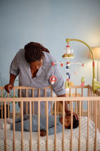 Dikey çekimde siyah anne yavaşça uyuyan bebeğe dokunuyor. Yatakta uzanmış, mavi duvarlı, rahat yatak odasında beşikten oyuncaklar asıyor.