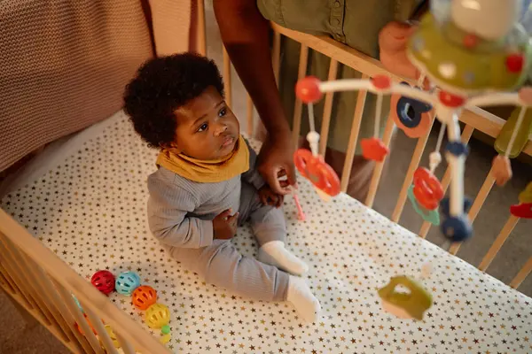 Yüksek açılı çekimde, yorgun siyah bebek yatakta oturur ve beşikteki hareketlere bakar. Annesi çocuğu sıcak bir ışıkla sıcak bir yatak odasında uyurken yukarıda asılı durur.