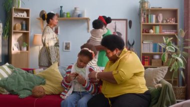 Afrikalı bir büyükanne torununa örgü örmeyi öğretirken, bir kadın, kucağında bebekle renkli bir oturma odasında kız kardeşiyle konuşuyor.