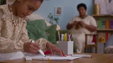 Keçeli kalemlerle çizim yapan, kahve masasında oturan ve büyükannesinin örgü ördüğü Afrikalı Amerikalı kız.