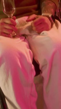 Doğumgünü partisinde kameraya bakıp şampanya kadehini kaldıran, arka planda parlak folyo perdesi ve balonlarla engelli, neşeli, beyaz bir adamın dikey portresi.