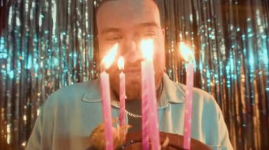 Mutlu beyaz bir adamın doğum günü pastasına mum üfleyip dilek tutarken, parlak folyo perdeyle süslenmiş duvarın karşısında dikilirken yakın plan çekimleri.