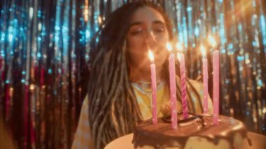 Kafkasyalı genç bir kadının doğum günü pastasına mum üflerken, kameraya gülümserken, alüminyum folyo perdelere yaslanırken, orta ölçekli yavaş çekimi.