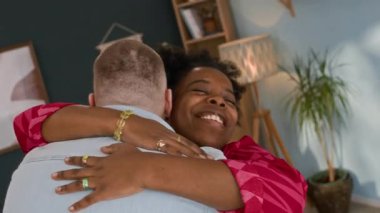 Orta ölçekli, gülümseyen, afro-amerikalı bir kadının beyaz erkek arkadaşına sarıldığı, evinde rahat bir oturma odasında buluştuğu ve konuştuğu bir fotoğraf.