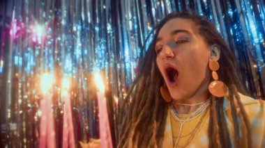 Kafkasyalı genç bir kadının doğum günü pastasına mum üflerken, parlak folyo folyo perdelerde otururken, doğum günü partisi yaparken yan görüntüsü.