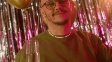 Kameraya bakıp dans eden beyaz bir adamın portresi şampanya kadehini kaldırıyor, arka planda folyo perdesi ve balonlar var.