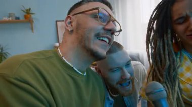 Yakın çekimde, mutlu beyaz arkadaşların favori karaoke şarkılarını mikrofona söylerken, evde rahat bir oturma odasında birlikte eğlenirken.