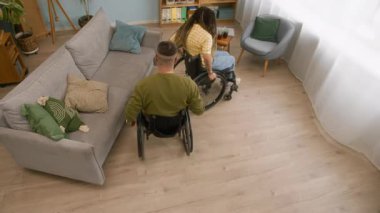 Yüksek açılı, yavaş çekim engelli beyaz arkadaşları tekerlekli sandalyelerle daireler çizip birbirlerini yakalamaya çalışıyorlar, gülüyorlar ve evde eğleniyorlar.