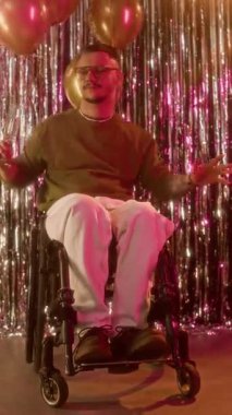 Engelli bir gencin dans ederken kameraya bakıp arka planda balonlarla şampanya kadehini kaldırmasının dikey portresi.