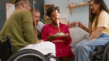 Mutlu, genç siyahi bir kadının doğum günü pastasının önünde parlak bir mumla dilek tutarken, doğum gününü evinde, tekerlekli sandalyeli yakın Kafkasyalı arkadaşlarıyla birlikte kutlarken orta uzun, yavaş bir çekim.