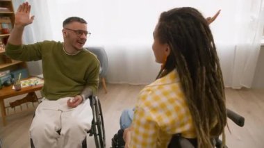 Yan görüş, tekerlekli sandalyedeki neşeli genç arkadaşların birbirlerine beşlik çakıp yumruk tokuşturarak modern oturma odasında birlikte vakit geçirmelerinin yavaş çekimi.