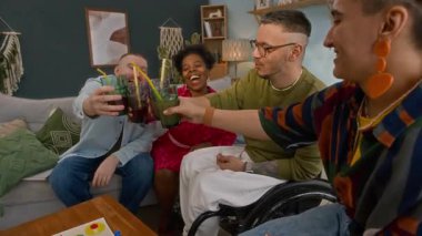 Neşeli çeşitli arkadaşlar ve tekerlekli sandalye kullanıcılarının limonata bardaklarını tokuşturup, gülüp, rahat bir oturma odasında keyif çattıkları, birlikte vakit geçirmekten mutlu bir çekim.