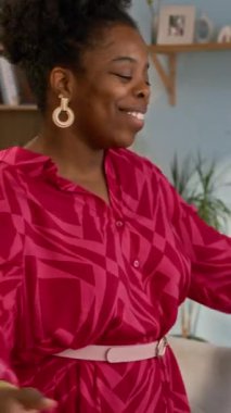 Süslü elbiseli, gülümseyen Afrikalı Amerikalı genç bir kadının tekerlekli sandalyedeki beyaz erkek arkadaşına sımsıkı sarılmasının, sıcak oturma odasında buluşmasının ve birbirimizi gördüğümüze sevinmesinin dikey görüntüsü.