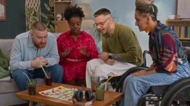 Mutlu genç arkadaşlar ve tekerlekli sandalye kullanıcılarından oluşan el kamerasıyla çekilmiş bir fotoğraf. Masa oyunları oynarken gülüyorlar, rahat bir oturma odasında oturuyorlar.