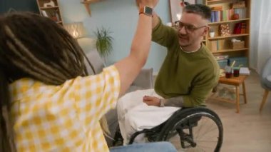 Tekerlekli sandalyedeki genç beyaz adamın önünde rastalı saçı olan ve modern oturma odasında eğlenen kadın arkadaşına beşlik ve yumruk tokuşturuşunun yüksek açılı yavaş çekimi.