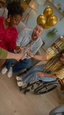 Oturma odasındaki koltukta oturan, tekerlekli sandalyeli arkadaşlarıyla şampanya bardaklarını tokuşturup, evlerinde eğlenen genç çiftin dikey yavaş çekimi.