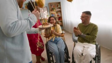 Çok ırklı mutlu genç arkadaşların tekerlekli sandalyedeki beyaz kadına doğum günü sürprizi yaparken çekilmiş bir fotoğraf. Yanında mumla doğum günü pastası getirip birlikte kutluyorlar.