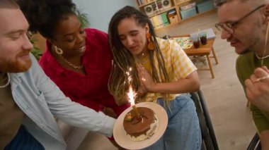 Tekerlekli sandalyede, doğum günü pastasının önünde mum ışığında dilek tutan, doğum gününü evinde çok ırklı arkadaşlarıyla birlikte kutlayan, mutlu, beyaz bir kadının yavaş çekimi.