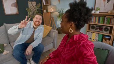 Gülümseyen genç siyahi kadın, neşeli beyaz erkek arkadaşıyla video bloğu kaydediyor rahat oturma odasında rahat bir ortamda oturuyor ve kameraya el sallıyor.