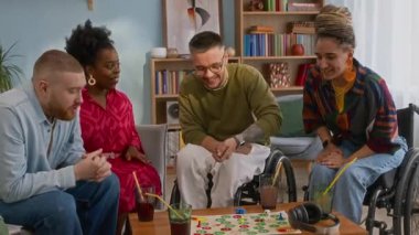 Çok ırklı genç arkadaşlar ve tekerlekli sandalye kullanıcılarının masa oyunları oynarken eğlendikleri, evlerinde rahat bir oturma odasında vakit geçirdikleri orta ölçekli bir fotoğraf.