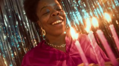 Doğum günü pastasında dilek tutan ve mum üfleyerek partideki parlak folyo perde duvarına karşı duran mutlu Afrikalı Amerikalı kadının yavaş çekimini büyüt.