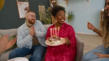 Mutlu genç siyahi kadının doğum günü pastasının önünde dilek tutarken ve doğum gününü kutlamak için tüm mumları üflerken yavaş çekimde yakınlaş tekerlekli sandalyeli Kafkasyalı arkadaşlarla.