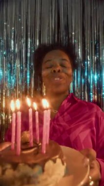 Mutlu genç siyahi kadının doğum günü pastasında dilek dileyip mum üflediği dikey zoom çekimi, partideki parlak folyo folyo perde duvarının karşısında duruyor.
