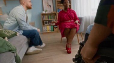 Tekerlekli sandalyedeki rahat oturma odasına giren beyaz bir kadının görüntüsü. Gülümseyen etnik çeşitlilikteki arkadaşlarına katılıyor. Birbirlerini görüp beşlik çakıyor.